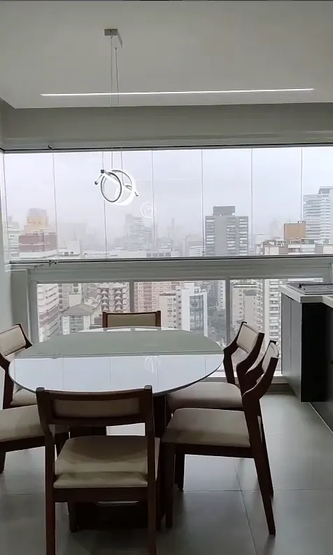 Mesa de jantar redonda com cadeiras de madeira diante da varanda envidraçada com vista para prédios